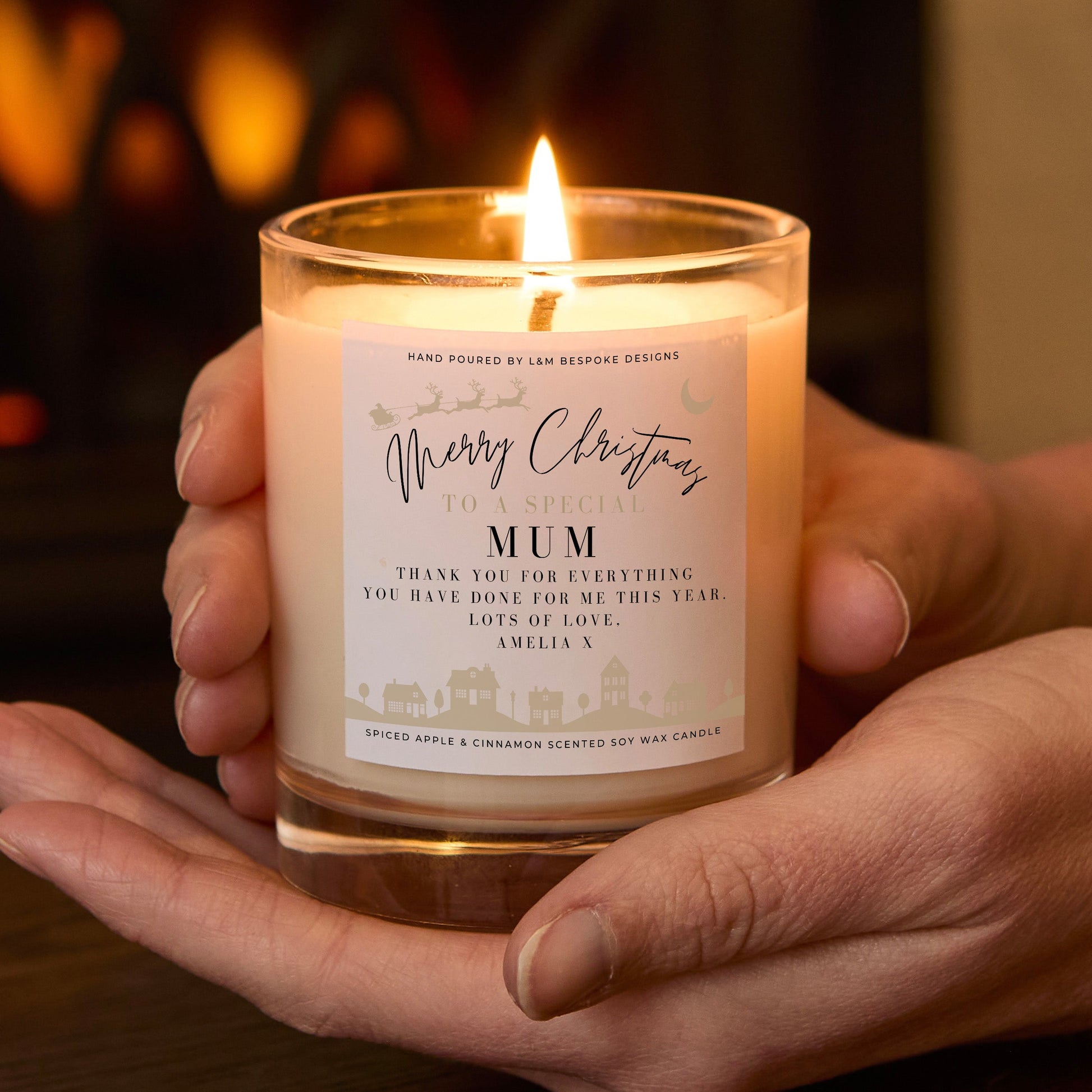 Hand holding a lit candle with a 'Merry Christmas Mum' label against a warm, blurred background.